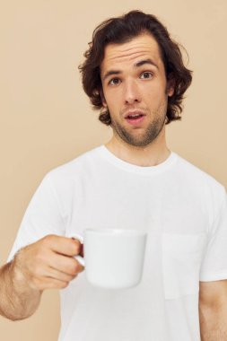 Ellerinde beyaz bir kupayla neşeli bir adam bej arka plan pozu veren duyguları var.