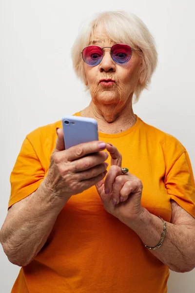 스마트폰을 손에 들고 감정을 확 잡고 유행하는 안경을 쓰고 있는 나이든 친절 한 여성의 모습 — 스톡 사진
