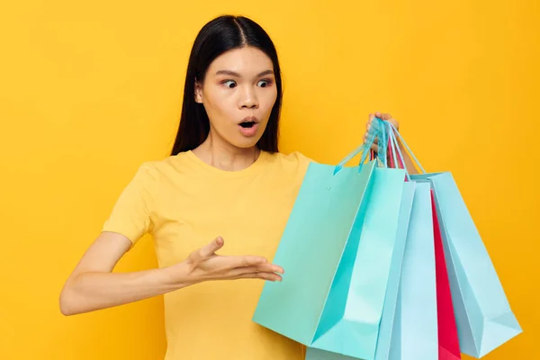 Charmante jonge Aziatische vrouw in een geel T-shirt met veelkleurige boodschappentassen gele achtergrond ongewijzigd — Stockfoto