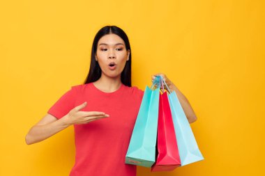Renkli çantalarıyla yakışıklı genç Asyalı kadın alışveriş pozu veriyor. Sarı arka plan değişmemiş.