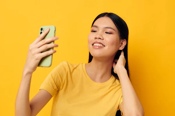 노란 티셔츠를 입고 휴대폰 포즈 스튜디오 모델을 보고 있는 아시아 출신의 여성 — 스톡 사진