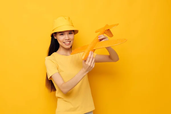 年轻的亚洲女人戴着一顶帽子，手里拿着一架飞机，迷人极了。玩具黄色背景不变 — 图库照片