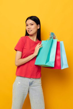 Renkli çantalarıyla yakışıklı genç Asyalı kadın alışveriş pozu veriyor. Sarı arka plan değişmemiş.