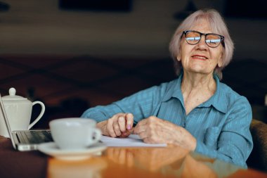 Bir fincan kahve ve dizüstü bilgisayarla bir kafede oturan yaşlı kadın.