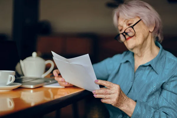 Mujer mayor con gafas se sienta en una mesa delante de un ordenador portátil Freelancer trabaja sin alteraciones — Foto de Stock
