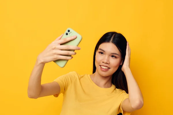 Affascinante giovane donna asiatica in una t-shirt gialla guardando il telefono posa sfondo isolato inalterato — Foto Stock