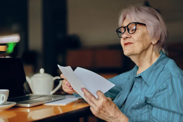 Mujer mayor con gafas se sienta en una mesa delante de un ordenador portátil Freelancer trabaja sin alteraciones — Foto de Stock