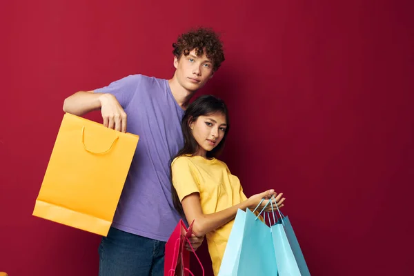 빨간 가방을 든 화려 한 티셔츠를 입은 귀여운 젊은 커플의 변화없는 빨간 배경 쇼핑 — 스톡 사진