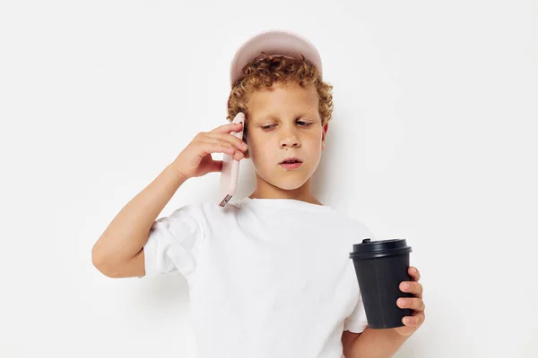 Lindo niño pequeño con una gorra de camiseta blanca con un teléfono en un vaso con un fondo claro bebida inalterado — Foto de Stock