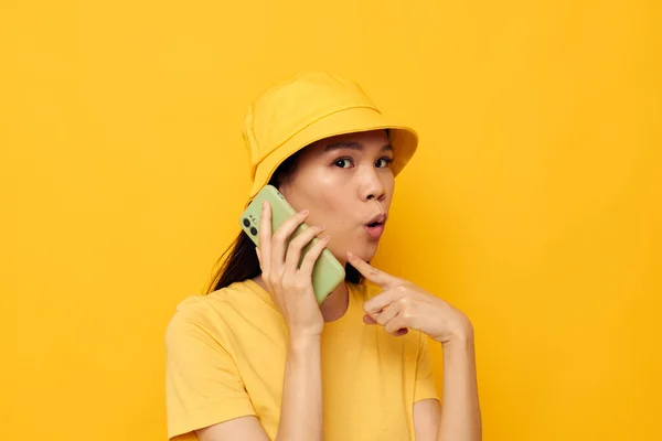 Affascinante giovane donna asiatica in un cappello giallo e T-shirt parlando sul modello di studio di telefono inalterato — Foto Stock