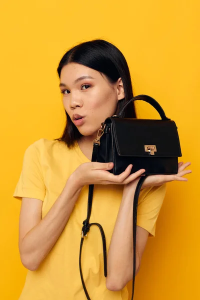 Funny asian woman in a yellow t-shirt black womens bag in hands posing — Stockfoto