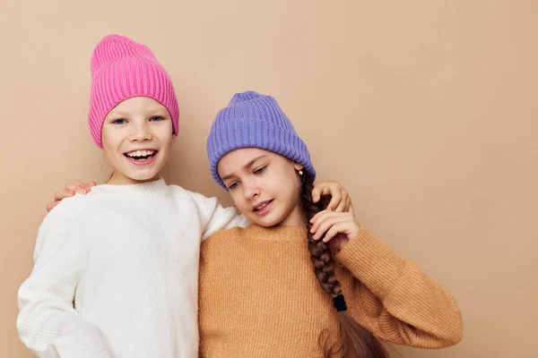 Две маленькие девочки в свитерах и шляпах обнимают бежевый фон дружбы — стоковое фото