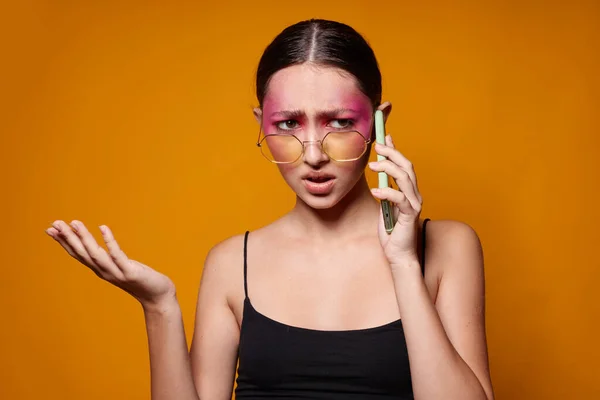 Vacker kvinna med telefon rosa ansikte makeup poserar attraktiva utseende glasögon gul bakgrund oförändrad — Stockfoto