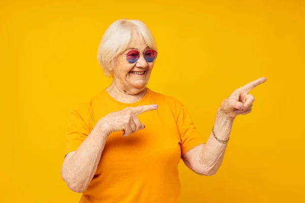 Portrait of an old friendly woman in casual t-shirt sunglasses yellow background — Stock Photo, Image