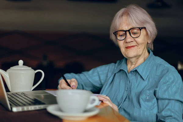 Hermosa anciana madura sentada en un café con una taza de café y un portátil Freelancer funciona sin cambios — Foto de Stock