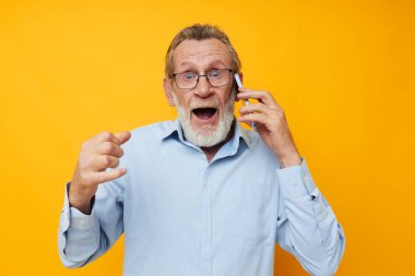 Mavi gömlekli ve gözlüklü yaşlı bir adam telefonda sarı arka planda konuşuyor.