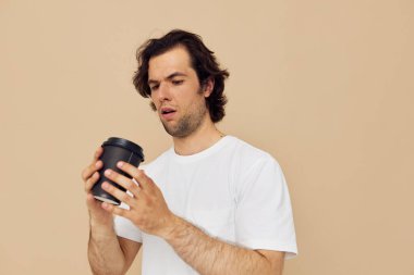Beyaz tişörtlü, elinde siyah bardakla bej arka planda neşeli bir adam.