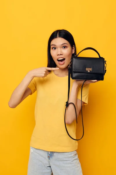 Funny asian woman in a yellow t-shirt black womens bag in hands posing — Stockfoto