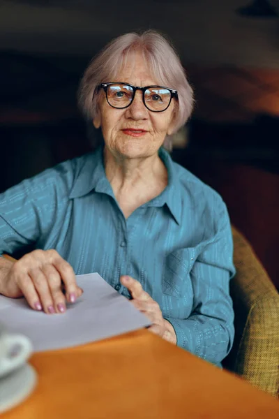Όμορφη ώριμη ηλικιωμένη γυναίκα με γυαλιά κάθεται σε ένα τραπέζι μπροστά από ένα φορητό υπολογιστή Κοινωνικά δίκτυα αναλλοίωτα — Φωτογραφία Αρχείου