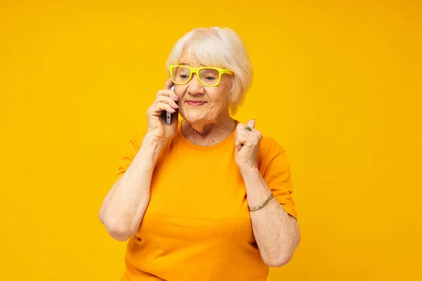 Портрет старой дружелюбной женщины разговаривающей по телефону в желтых очках на изолированном фоне — стоковое фото