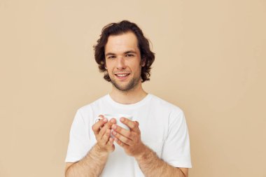 Ellerinde beyaz bir kupayla neşeli bir adam bej arka plan pozu veren duyguları var.