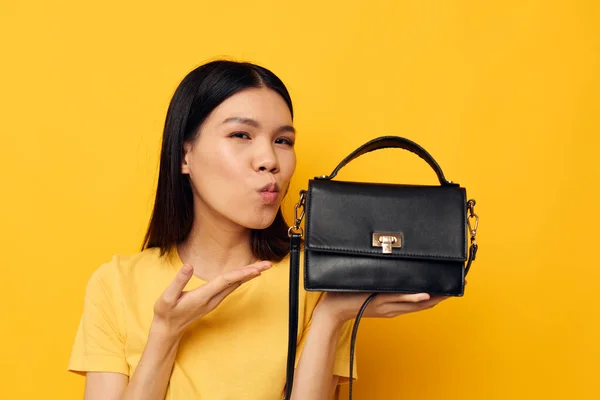 Rolig asiatisk kvinna i en gul t-shirt svart kvinnors väska i händerna poserar — Stockfoto
