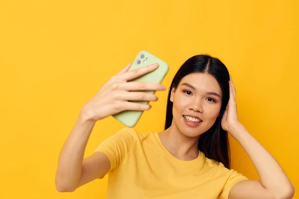Γοητευτική νεαρή Ασιάτισσα με ένα κίτρινο μπλουζάκι κοιτάζοντας το τηλέφωνο με απομονωμένο φόντο αναλλοίωτο. — Φωτογραφία Αρχείου