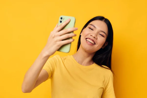 노란 티셔츠를 입고 휴대폰 포즈 스튜디오 모델을 보고 있는 아시아 출신의 여성 — 스톡 사진