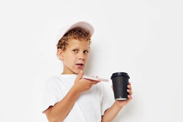 Foto retrato niño rizado en una gorra de camiseta blanca con un teléfono en un vaso con un fondo claro bebida inalterado — Foto de Stock
