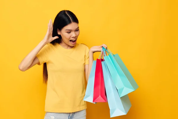 Charmante jonge Aziatische vrouw in een geel T-shirt met veelkleurige boodschappentassen gele achtergrond ongewijzigd — Stockfoto