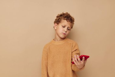 Elinde telefon olan kazaklı bir çocuk bej rengi arka planda.