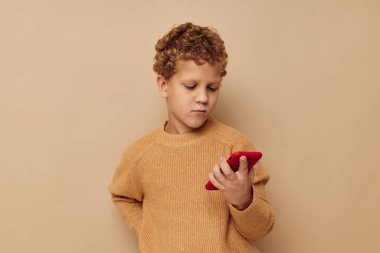 Elinde telefon olan kazaklı bir çocuk bej rengi arka planda.
