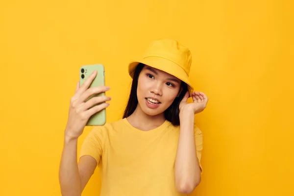 Donna con aspetto asiatico in un cappello giallo e T-shirt parlando sul telefono sfondo giallo inalterato — Foto Stock