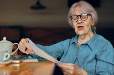 Gözlüklü yaşlı kadın dizüstü bilgisayarın önünde masada oturuyor. Freelancer değişmeden çalışıyor.
