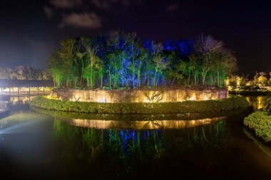 Aydınlatılmış beton setin üzerindeki ağaçların manzarası gölün su yüzeyine yansıyor ve arka planda gece köprüsü var.