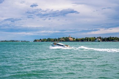 Sürat teknesi deniz suyu yüzeyinde hızla ilerlerken, arka planda bulutlu gökyüzüne karşı bina inşa ediliyor.