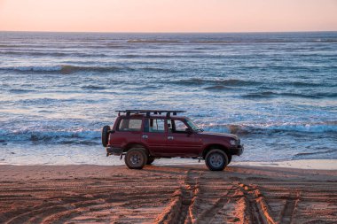 Alacakaranlıkta kumlu sahilde park etmiş kırmızı off-road arabası güzel deniz manzarasının önünde.
