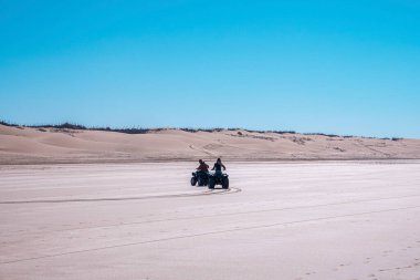 İnsanlar kum tepecikleriyle birlikte kumlu yolda dörtlü bisiklete biniyorlar.