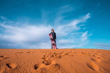 Man with sandboard standing on sand dunes in desert against cloudy sky