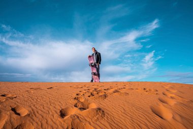 Man with sandboard standing on sand dunes in desert against cloudy sky