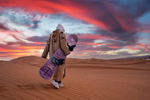Man in traditional clothes with sandboard walking on sand dunes against sky — Zdjęcie stockowe