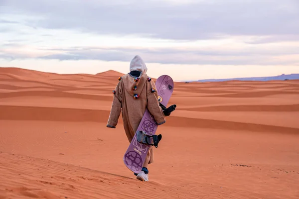 Man in traditional clothes with sandboard walking on sand dunes against sky — Foto Stock