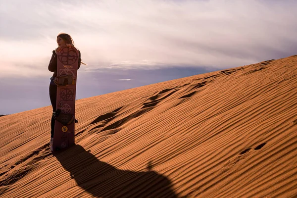 Woman with sandboard standing on sand dunes in desert against cloudy sky — Foto Stock