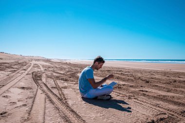 Man using laptop sitting on beach on a bright sunny day