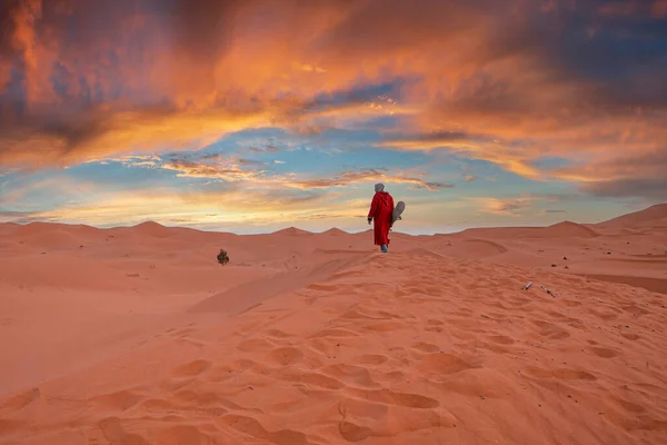 Man in traditional clothes with sandboard walking on sand dunes during dusk — Foto Stock