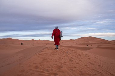 Man in traditional clothes with sandboard walking on sand dunes against sky