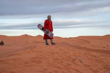 Man in traditional clothes with sandboard walking on sand dunes against sky