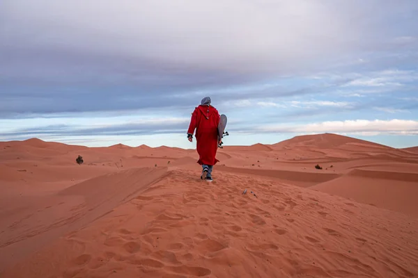 Man in traditional clothes with sandboard walking on sand dunes against sky — Zdjęcie stockowe