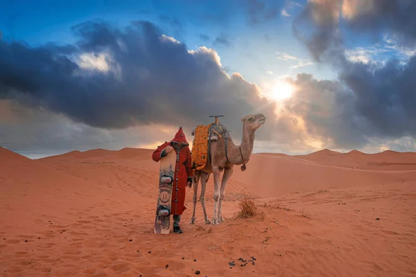 Man in traditional clothes with sandboard standing beside camel in desert — Zdjęcie stockowe