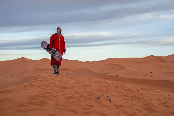 Man in traditional clothes with sandboard standing on sand dunes against sky — Foto Stock